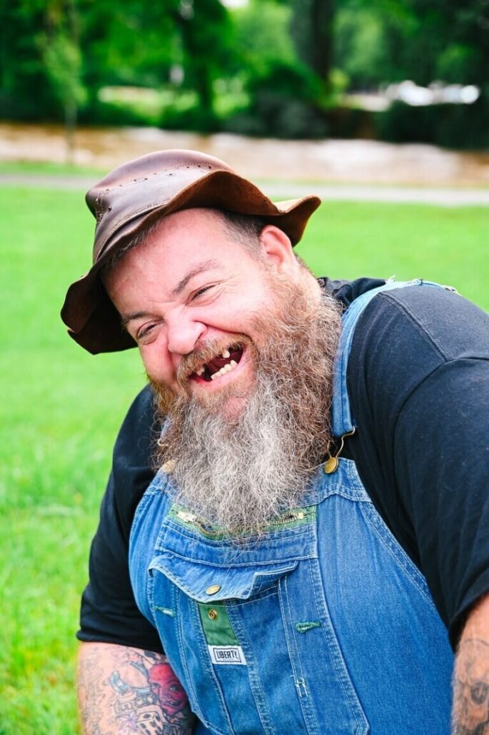 Theredneckbus Tour Smiling Man Beard Hat Grass